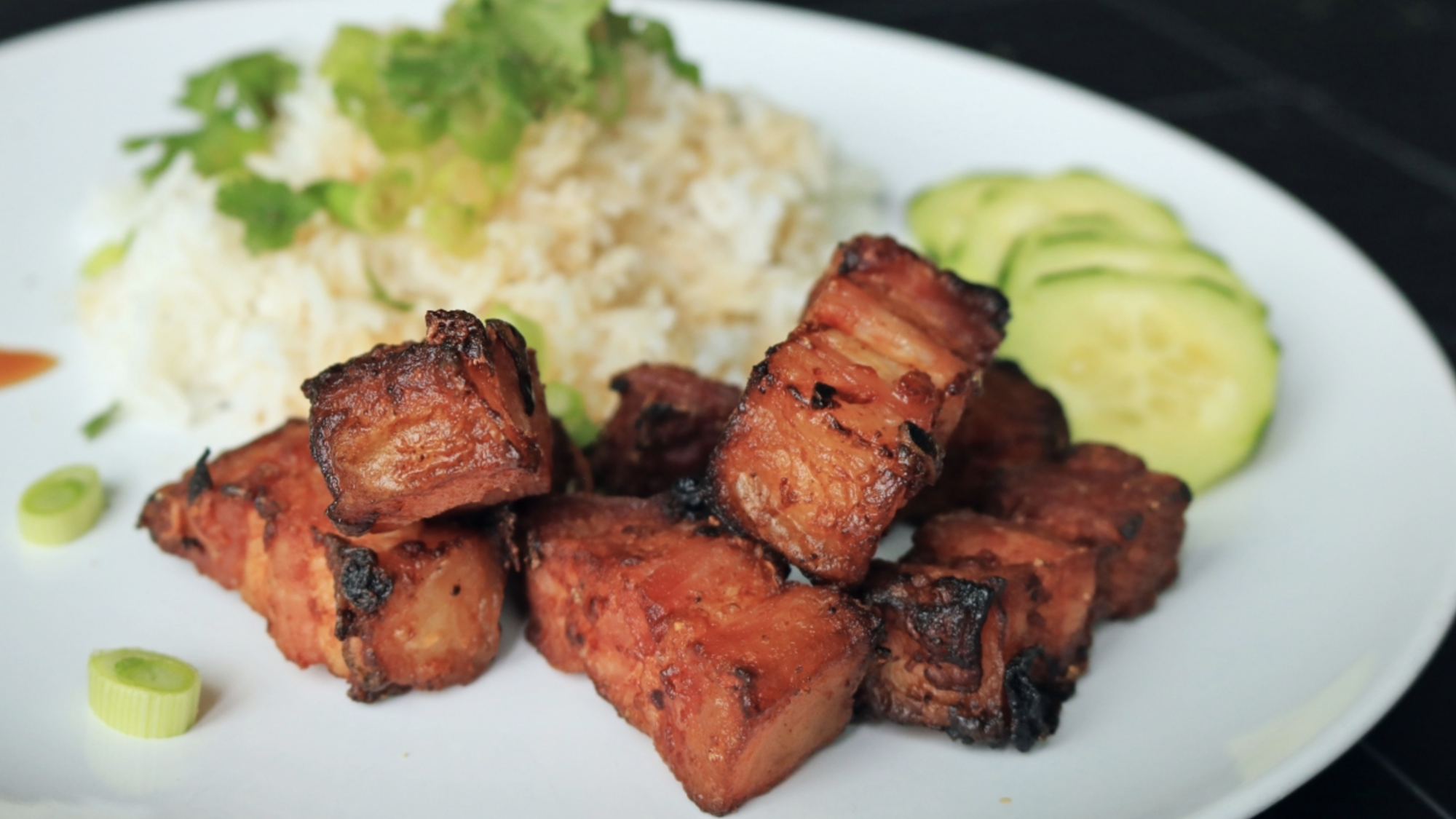 Air fried pork belly pieces on a plate with rice and cucumbers.