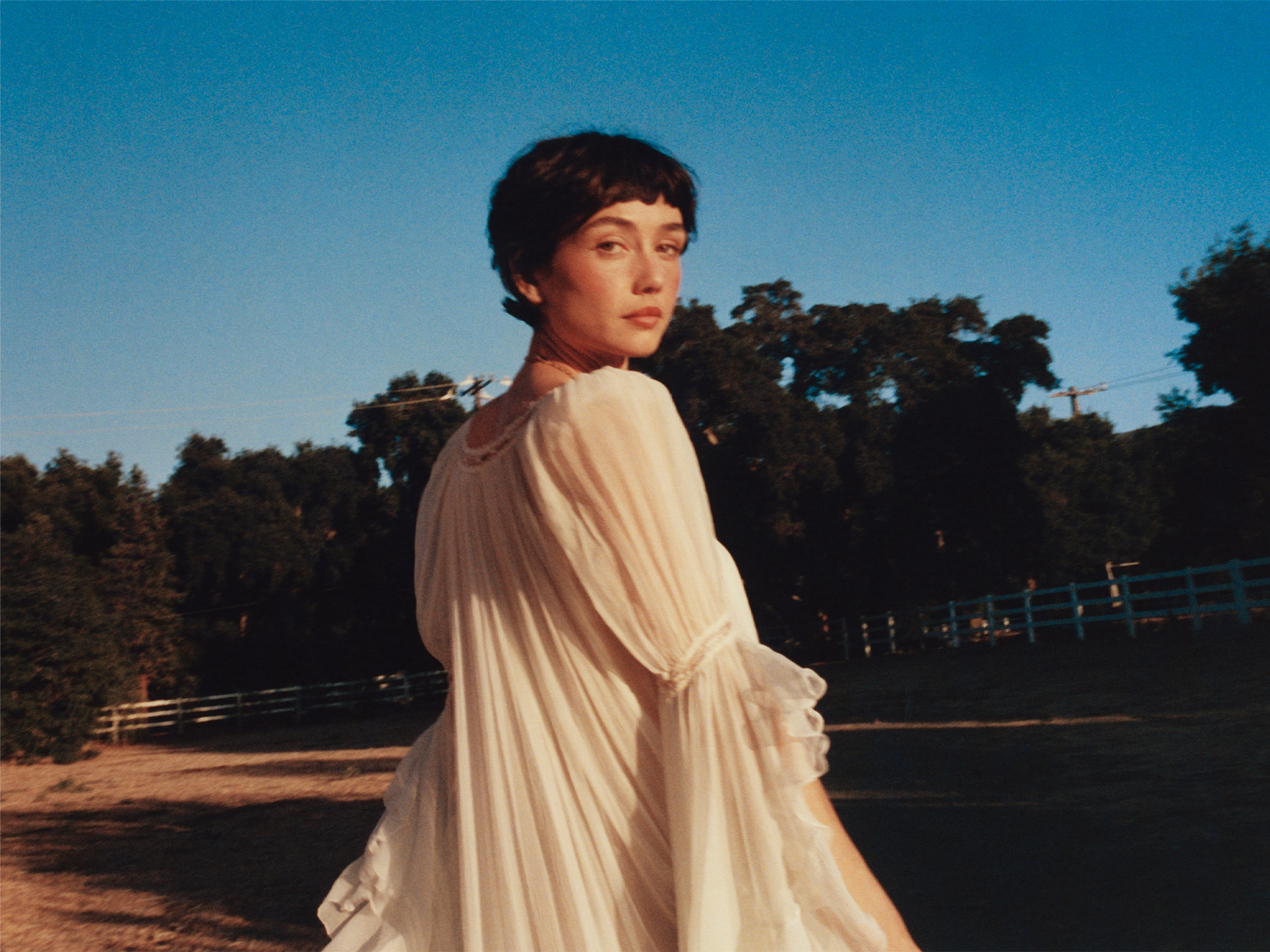 Sydney Chandler looks back at the camera. She is on a ranch and wearing a mint green Chloe ruffled dress.