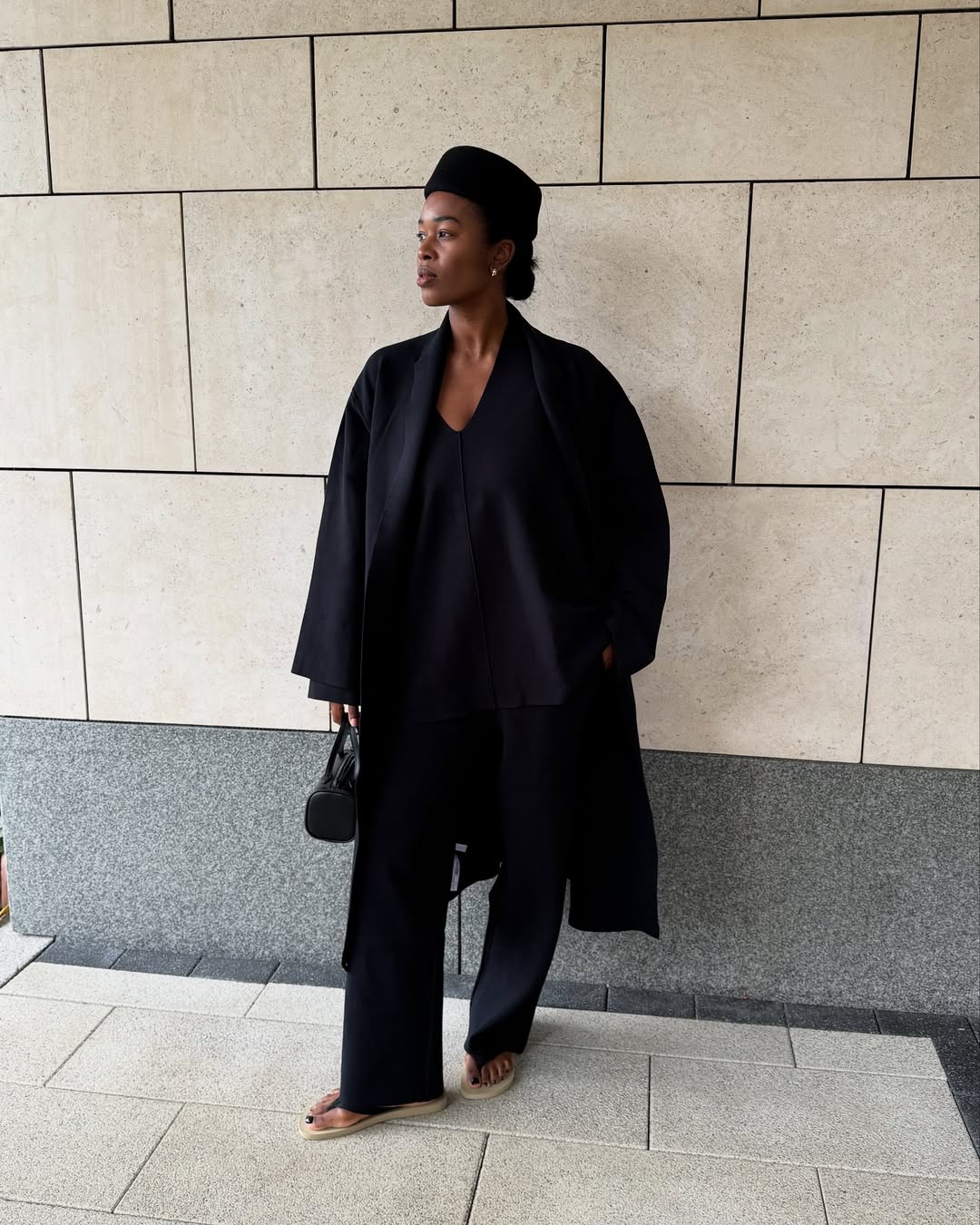 Content creator Marilyn NK wearing an all-black outfit consisting of a long coat, black trousers and a hat.