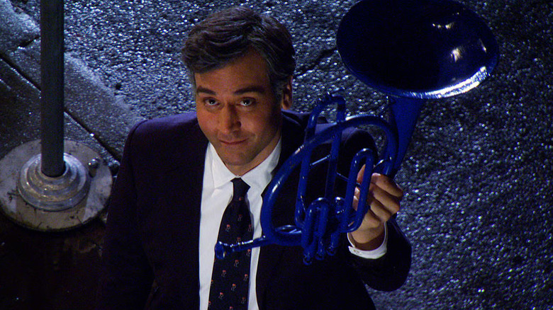 Ted smirks as he holds up a blue-painted French horn while standing beside a streetlamp pole with the dark road behind him in How I Met Your Mother